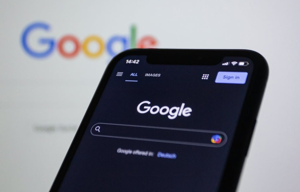 A close-up photograph of a smartphone displaying the Google search homepage in dark mode, positioned in front of a computer screen which shows a slightly out-of- focus version of the same colorful Google logo. The image highlights how the Google search interface appears on mobile devices.