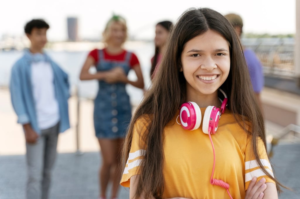 Portrait of cute smiling teenage girl wearing headphones, yellow t shirt looking at camera on the street with friends on background