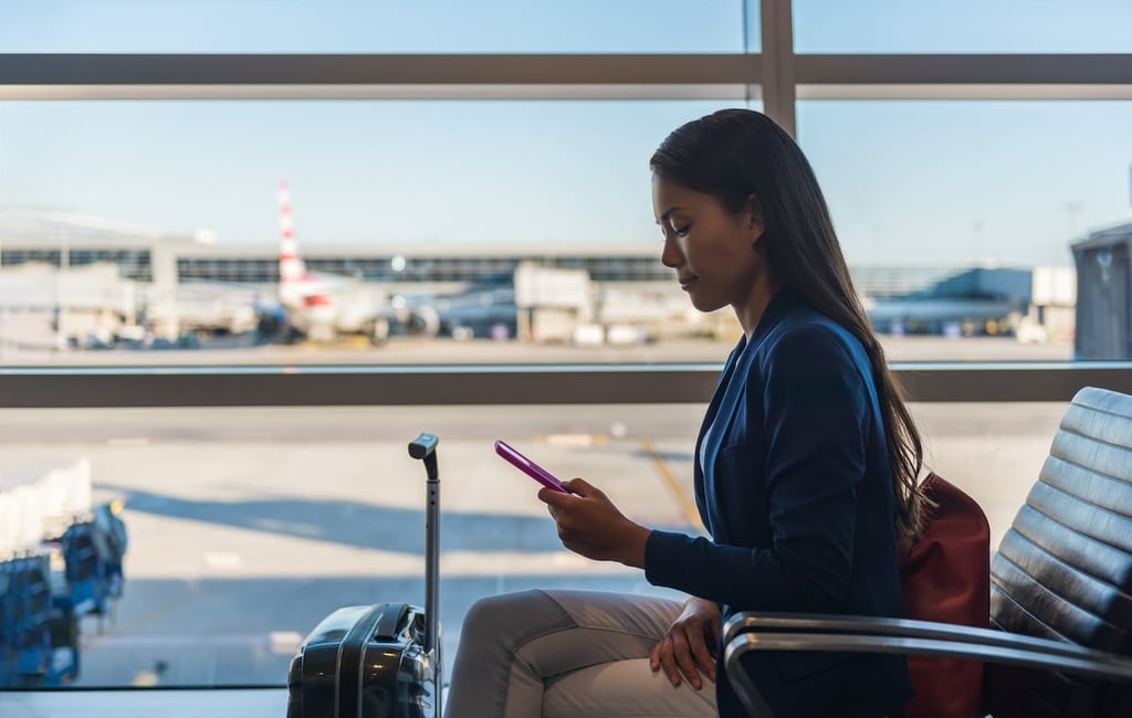 Stock photo of a traveler sitting at an airport terminal looking at her phone.