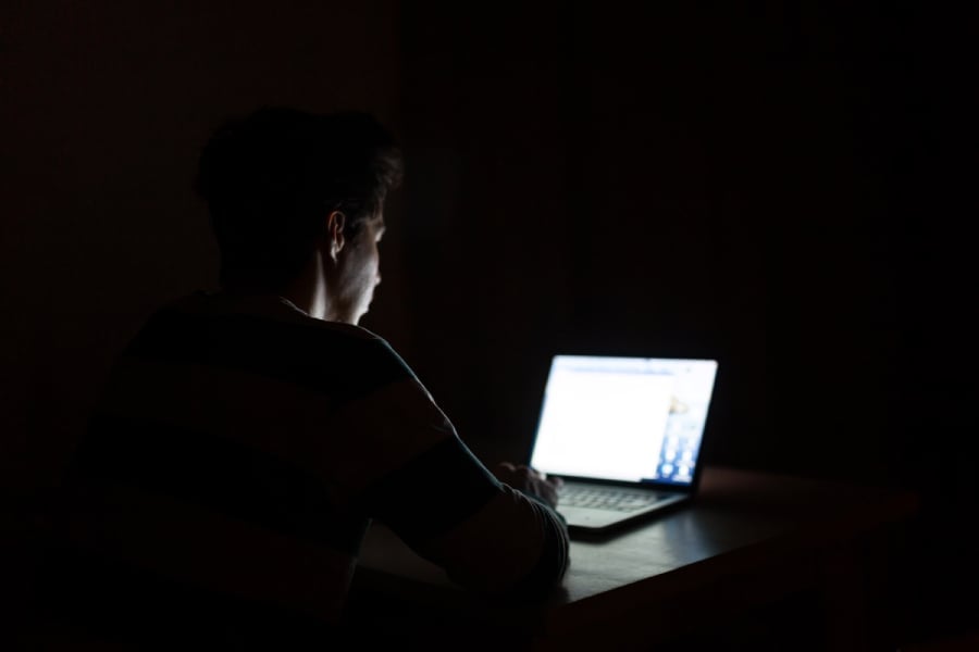 A man browsing through his laptop inside a dark room.