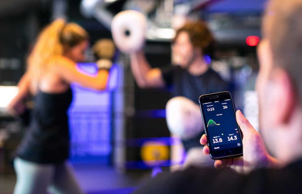 A person holds a smartphone displaying a fitness app with a graph and workout data in the foreground, while two other people are out of focus in the background, boxing in a gym.