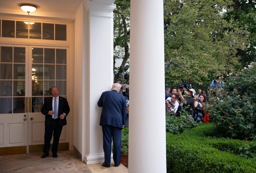 Donald Trump leans against a column outside the White House as members of the press gather with cameras and microphones.