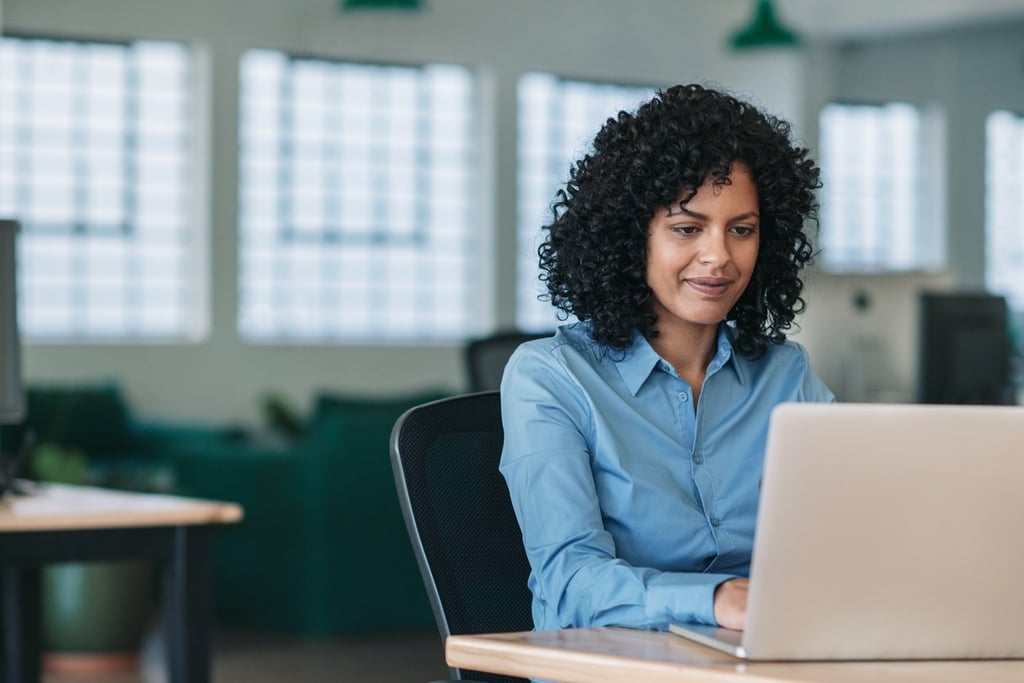 A smiling female employee with curly hair sitting at a desk and working on a laptop in a modern office.