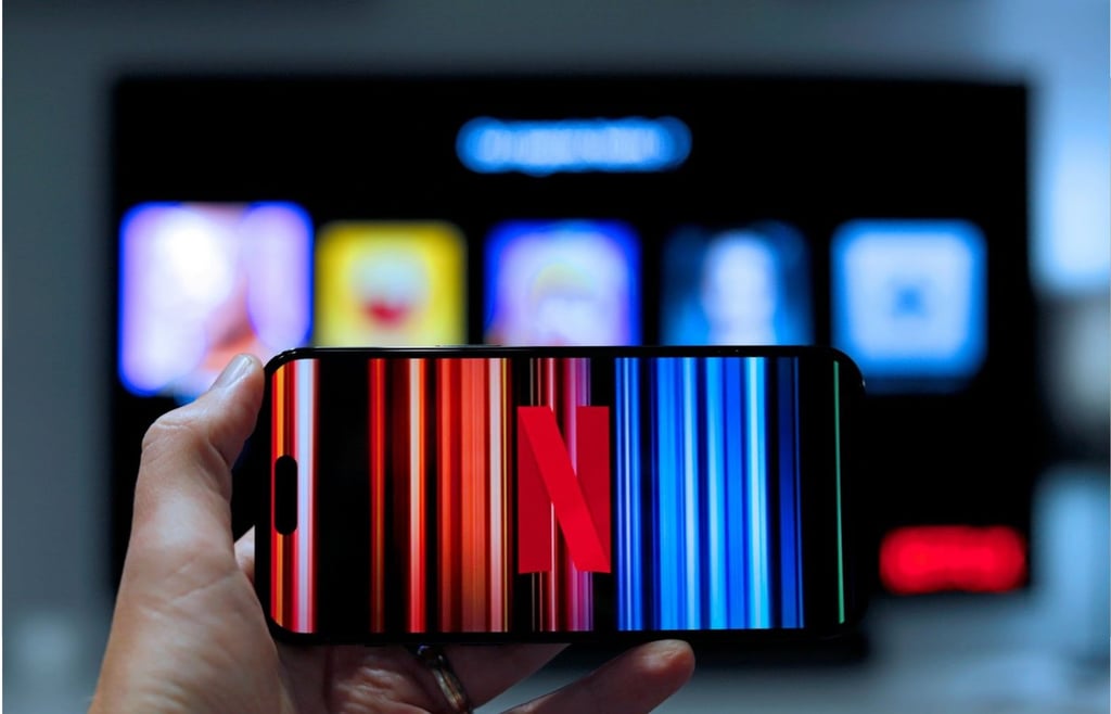 A hand holding a smartphone displaying the Netflix logo and colorful light streaks, with a blurred TV screen showing the Netflix app interface in the background.