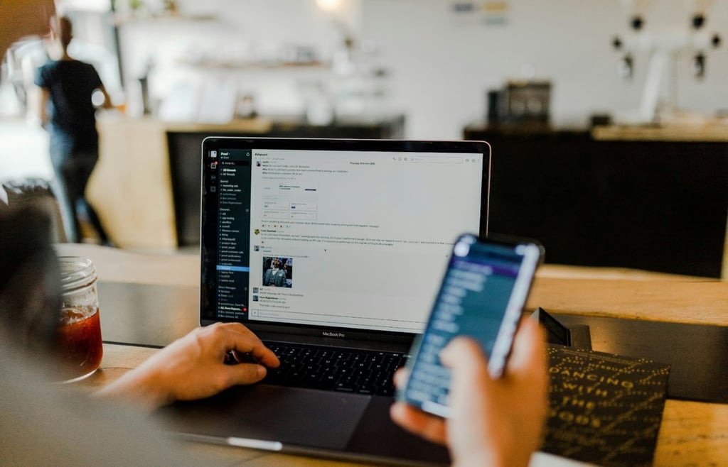 A person works on a laptop showing a Slack chat interface while holding a smartphone.