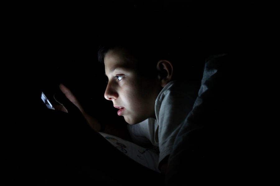 A young boy watching on his smartphone while on bed inside a dark room.