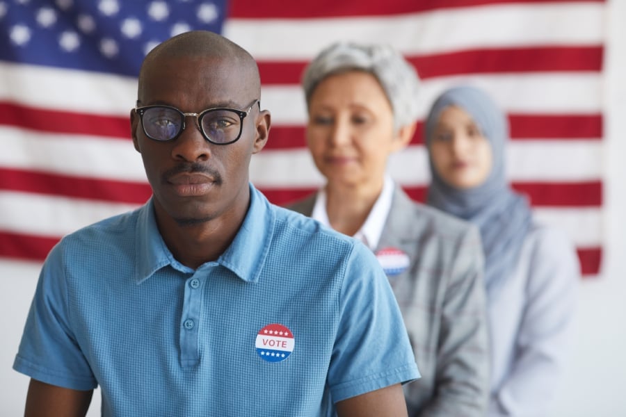 A small group of people with diverse ethnicity preparing to vote with the USA flag in the background.