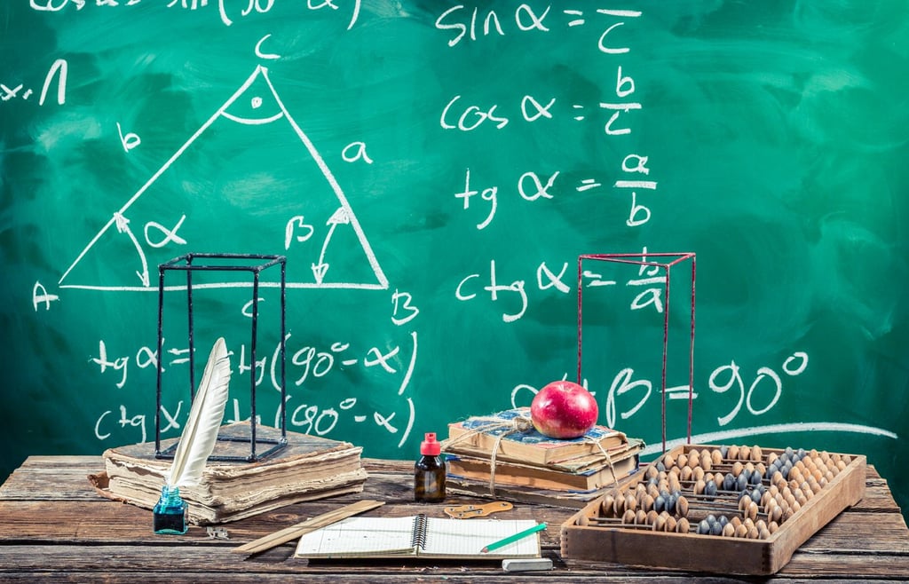 Mathematical implements on a wooden table with scribbled equations on a greenboard.