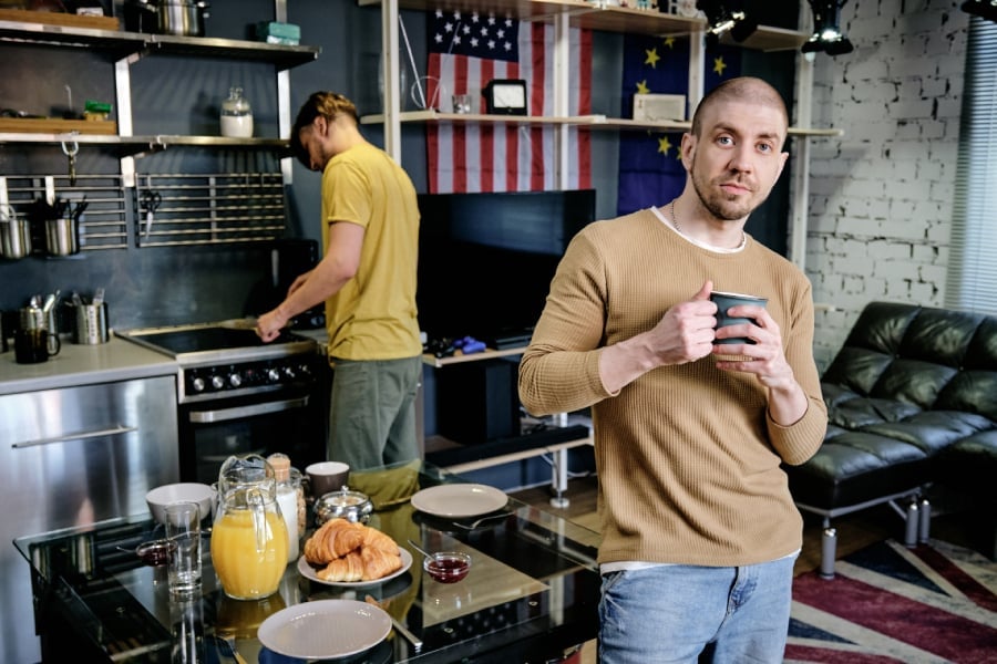 Young man with mug standing by table against another guy.