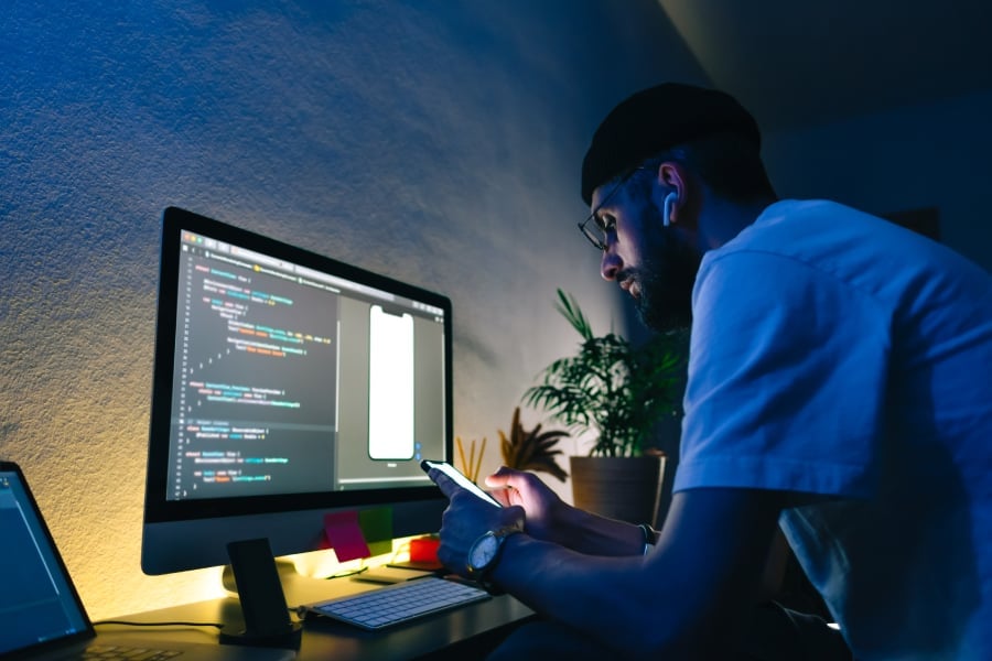 Young man mobile developer writes program code on a computer and check app in smartphone.