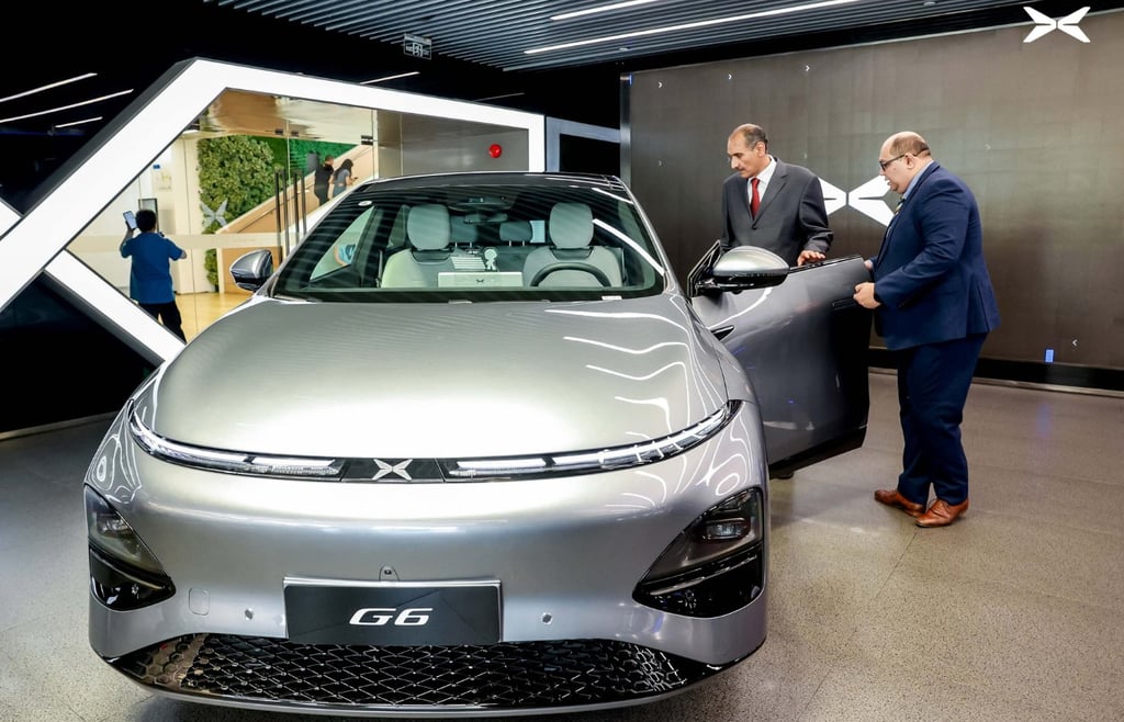 Silver XPeng G6 electric coupe SUV on display at a car showroom with two men looking at the open driver-side door.